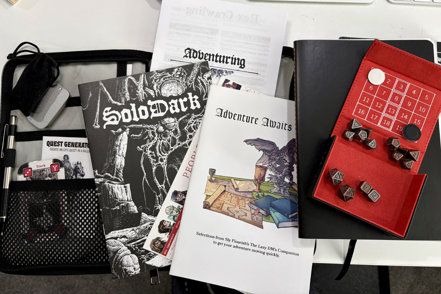 A tabletop role-playing game kit on a white desk, featuring a "SoloDark" rulebook, an "Adventure Awaits" booklet, a black notebook, and a red dice tray containing several dice.